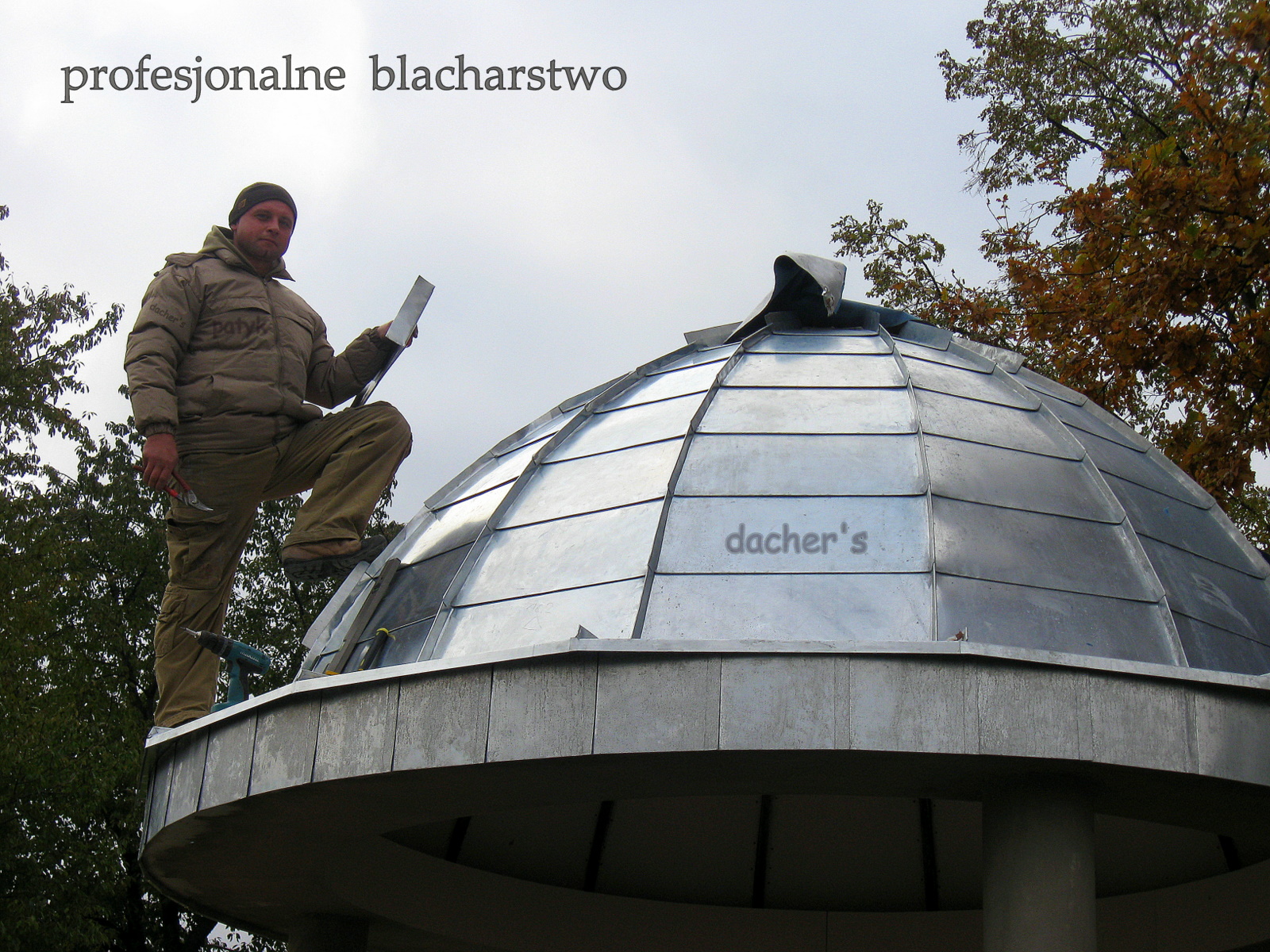 Blacharz w kurtce i czapce, stojący na kopule pokrytej blachą, trzymający płaski element blachy w ręku, w tle drzewa i pochmurne niebo.
